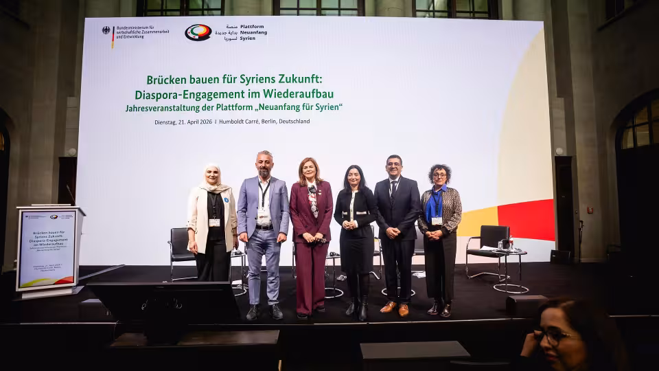 Fünf Personen stehen nebeneinander auf einer Bühne vor einer großen Leinwand mit Text zur Diaspora-Engagement-Veranstaltung für Syrien
