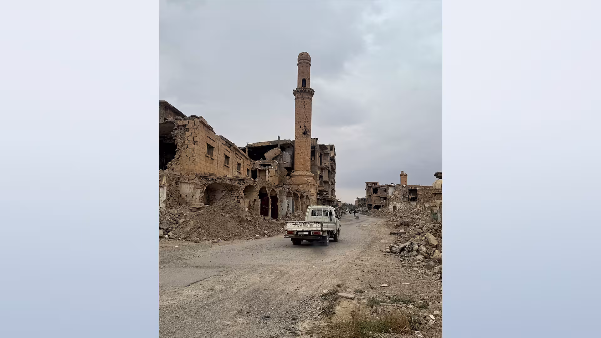 Eine Straße führt durch ein zerstörtes Stadtviertel mit beschädigtem Minarett und einem Kleintransporter.