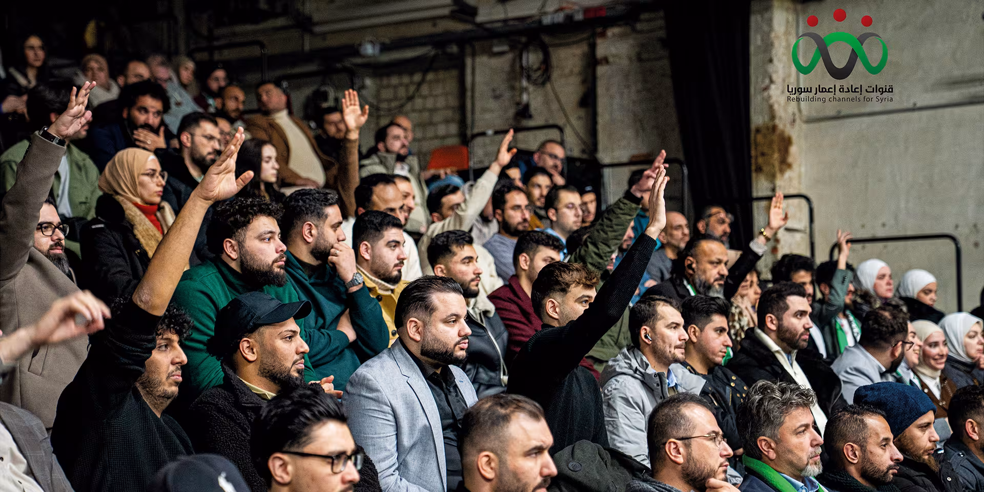 Teilnehmende einer Versammlung heben die Hände zur Abstimmung; oben rechts steht das Logo „Rebuilding Channels for Syria“.