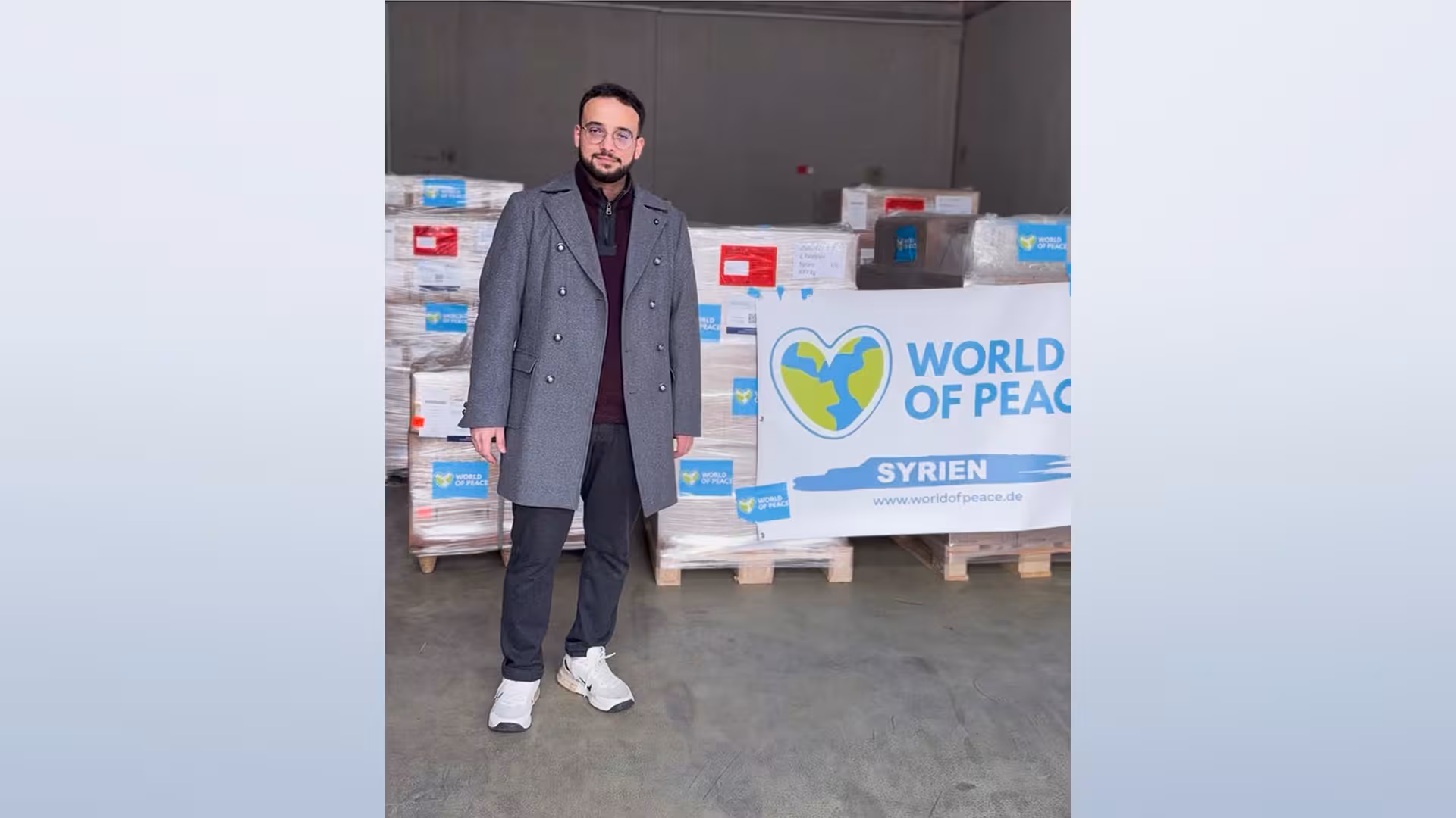 Ein Mann steht in einer Lagerhalle vor Hilfsgüterpaletten und einem Schild mit der Aufschrift „World of Peace – Syrien“.