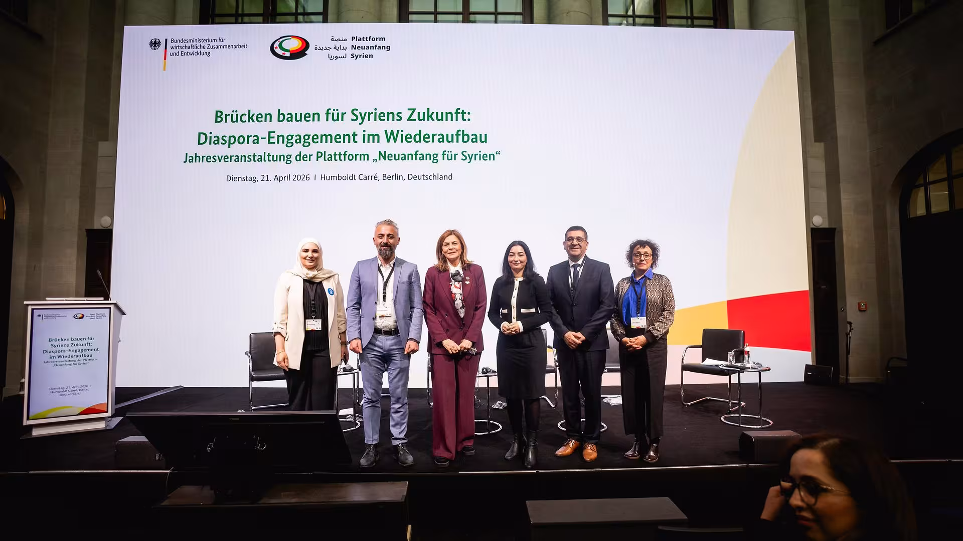 Fünf Personen stehen nebeneinander auf einer Bühne vor einer großen Leinwand mit Text zur Diaspora-Engagement-Veranstaltung für Syrien